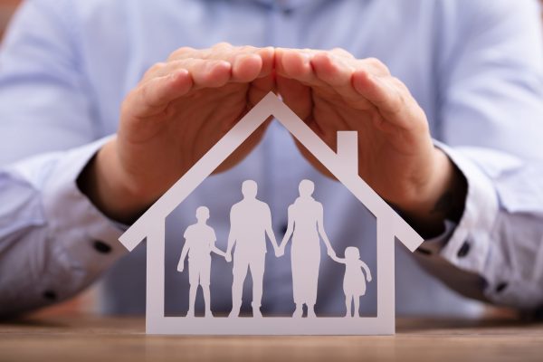 Close-up Of A Businessman Protecting Family Paper Cut Out With Hands On Wooden Desk